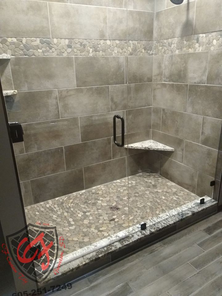 Gray large-format tile walk-in shower with pebble-stone floor, corner shelf and black-handled glass door